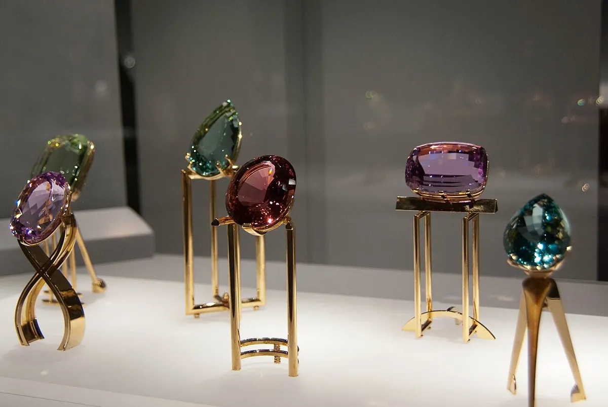 Large cut gemstones in red, purple, green and blue displayed on gold stands in a museum case