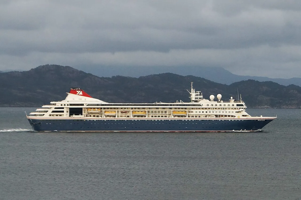 A cruise ship with a dark blue hull and a red funnel sailing through a fjord in front of mountains
