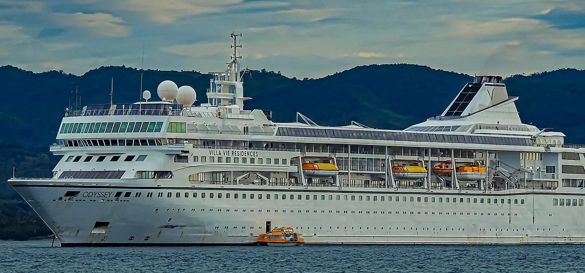 The white cruise ship Villa Vie Odyssey at anchor in front of green hills, with the name Odyssey on its bow