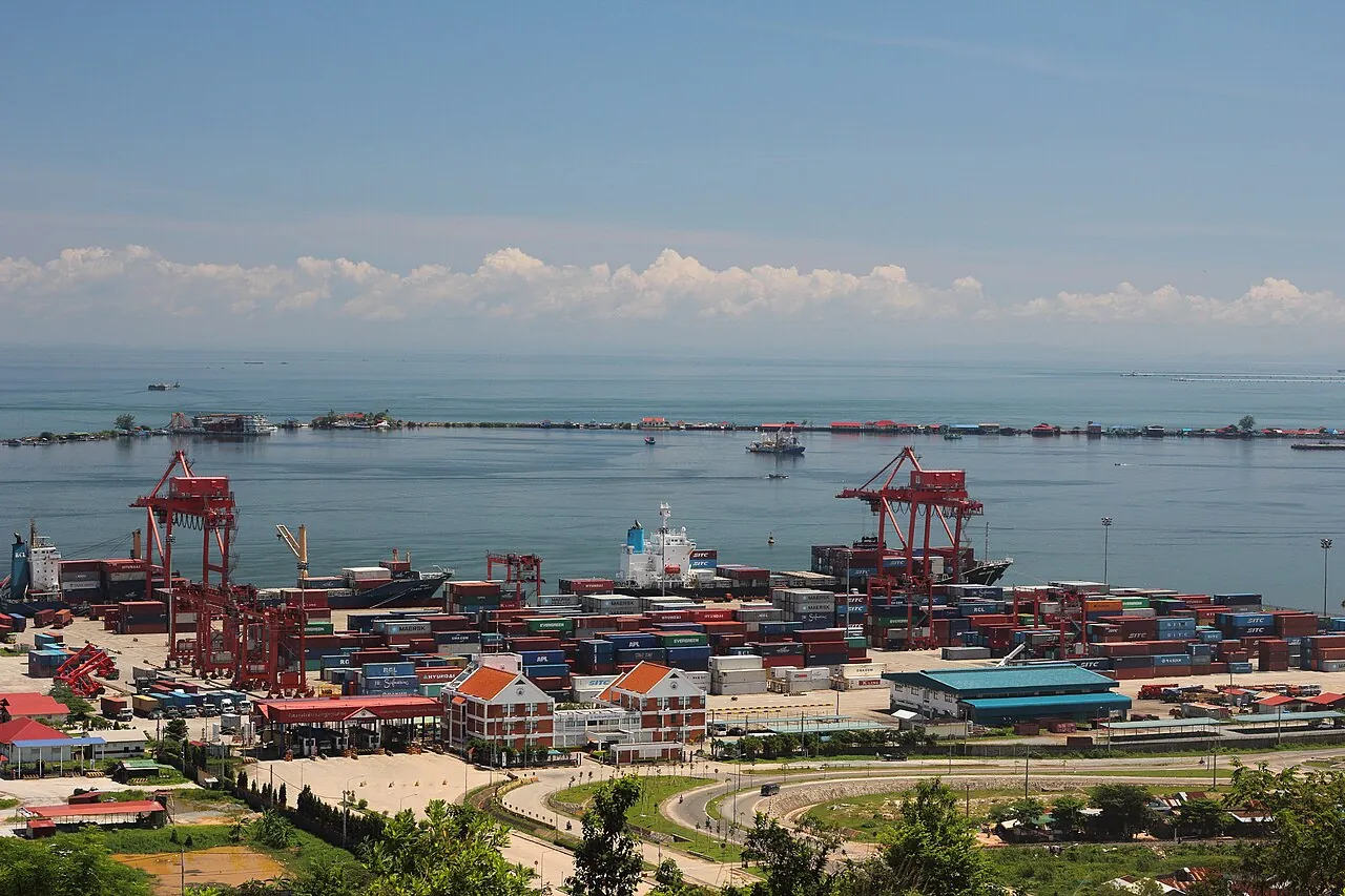 Red container cranes and stacks of shipping containers at Sihanoukville port beside the sea, seen from a hill